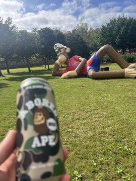 A hand holding a BAPE (Bathing Ape) branded can in the foreground, with a large statue of a BAPE character reclining on the grass in a park setting.