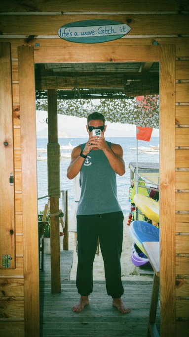 A man wearing sunglasses and a gray tank top takes a mirror selfie in a wooden beach hut doorway, with a sign above reading 'life's a wave...Catch it.' and an ocean view behind him.
