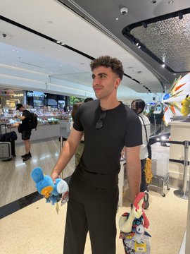 A young man poses at what appears to be a shopping mall, holding up plush toys including a blue Piplup, with the post indicating he's saying goodbye to Singapore while visiting a Pokémon Center before departure.