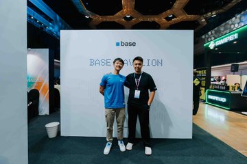 Two men pose together in front of a 'base' branded backdrop with 'BASE PAVILION' text, appearing to be at a conference or event.