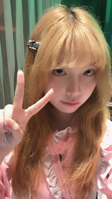 A close-up selfie of a young woman with long blonde hair and bangs, wearing a pink ruffled top and a sparkly hair clip, making a peace sign with her hand.