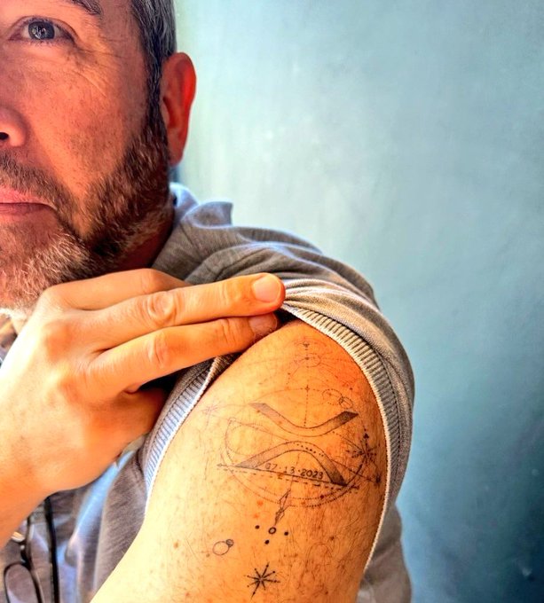 A man pulls down his sleeve to reveal a tattoo on his upper arm featuring a stylized "X" design with the date "22-11-2023" and small celestial elements, relating to the post about a CEO getting an XRP tattoo to mark his commitment to the cryptocurrency.