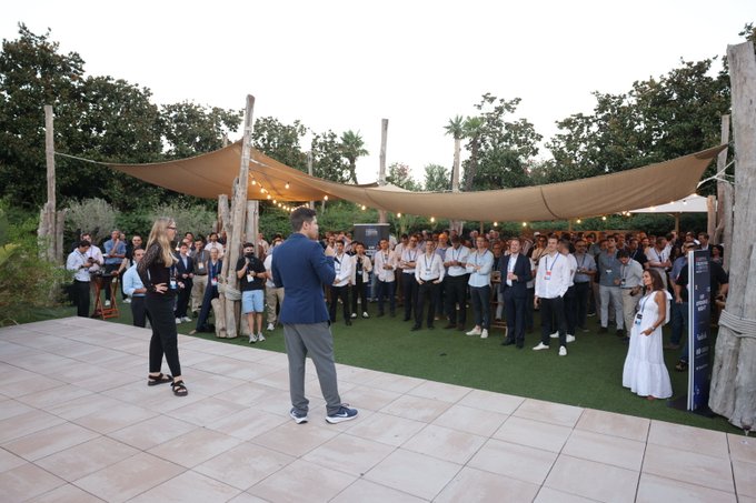 An outdoor evening event where a man in a blue jacket addresses a crowd gathered under large tan shade sails.