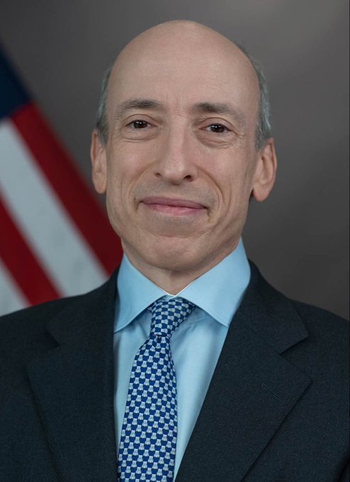 An official portrait of a bald man in a dark suit and blue checkered tie standing in front of an American flag.