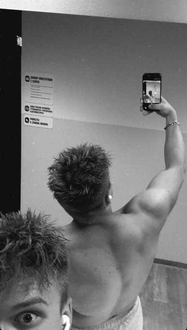 A black-and-white photo shows two shirtless men, with one in the foreground taking a mirror selfie with a smartphone in a gym, while the other peeks into the bottom of the frame. Wall signs read "EXHIBIT BEAUTY/MIRROR ETICONE" and "DO NOT WEAR SHOES/CLOTHES IN TRAINING".