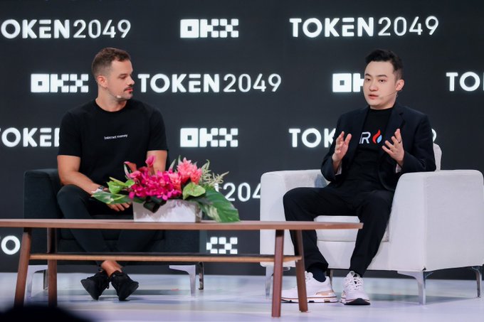 Two men sit on white chairs on a stage at the TOKEN2049 conference sponsored by OKX, with one speaking while gesturing with his hands.