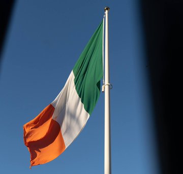 The image shows the Irish flag—a vertical tricolor of green, white, and orange—waving against a clear blue sky on a flagpole. It connects to the post by illustrating the country, Ireland, that has opted to follow the EU's guidance and exclude crypto from tax-advantaged savings accounts while still permitting related investment vehicles like crypto ETFs.