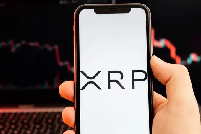 A hand holding a smartphone displaying the XRP logo on screen, with a blurred laptop keyboard and a red/green trading chart visible in the background.