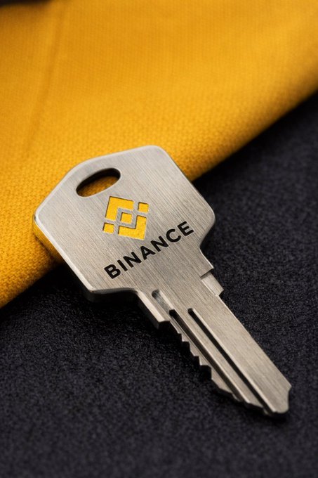 A silver key featuring the Binance logo rests on a yellow fabric against a dark textured surface.