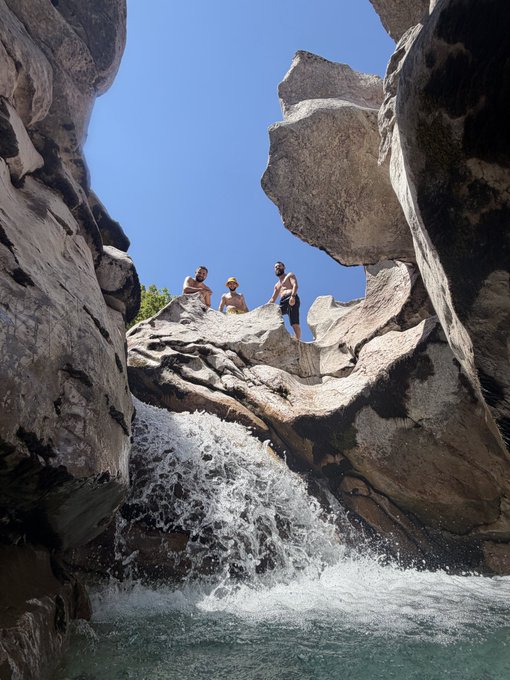 Three people stand on a rocky ledge above a cascading waterfall, framed by large boulders against a clear blue sky, suggesting a scenic natural getaway spot being shared with potential visitors from the crypto community. The image complements the post's invitation by showcasing the picturesque outdoor destination.