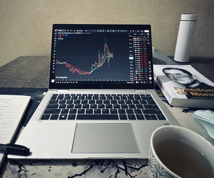 A single laptop displays a crypto trading chart beside a notebook, pen, coffee, and a book, reinforcing the message that you can trade crypto with just one laptop, one pen, and one diary.