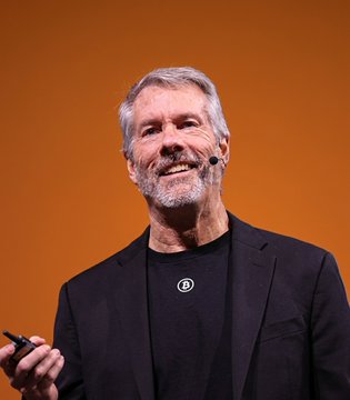 The image features a man wearing a black suit, standing against an orange background with his hands raised. He has a microphone attached to his face, suggesting that he might be giving a speech or presentation. The main focus of the image is on Michael Saylor, who appears to be smiling and engaging with the audience. In the bottom right corner of the image, there's a post that reads "Michael Saylor: 'Bitcoin keeps working'". This post seems to be related to the man speaking at the event or possibly discussing Bitcoin in general.