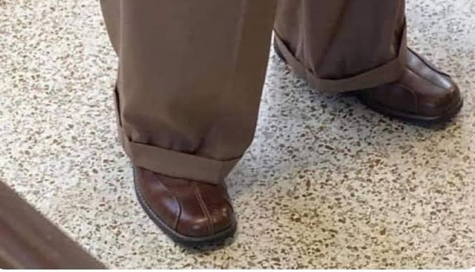 A close-up of brown leather shoes and cuffed brown trousers standing on a speckled terrazzo floor.