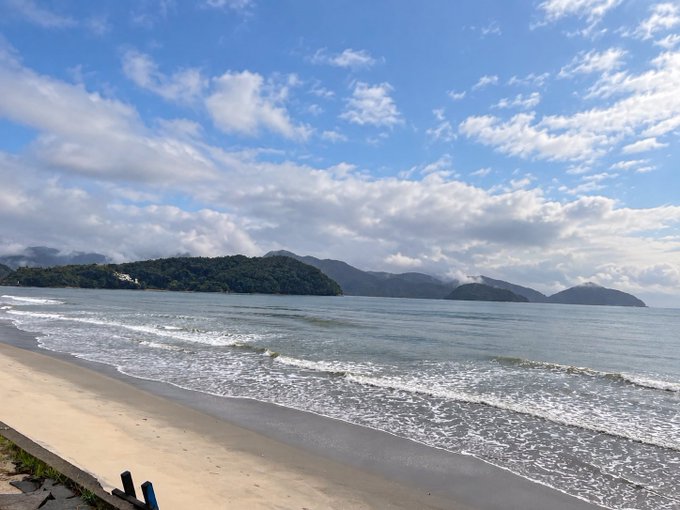 A scenic view of a beach with gentle waves, sandy shore, and forested mountains under a partly cloudy blue sky.