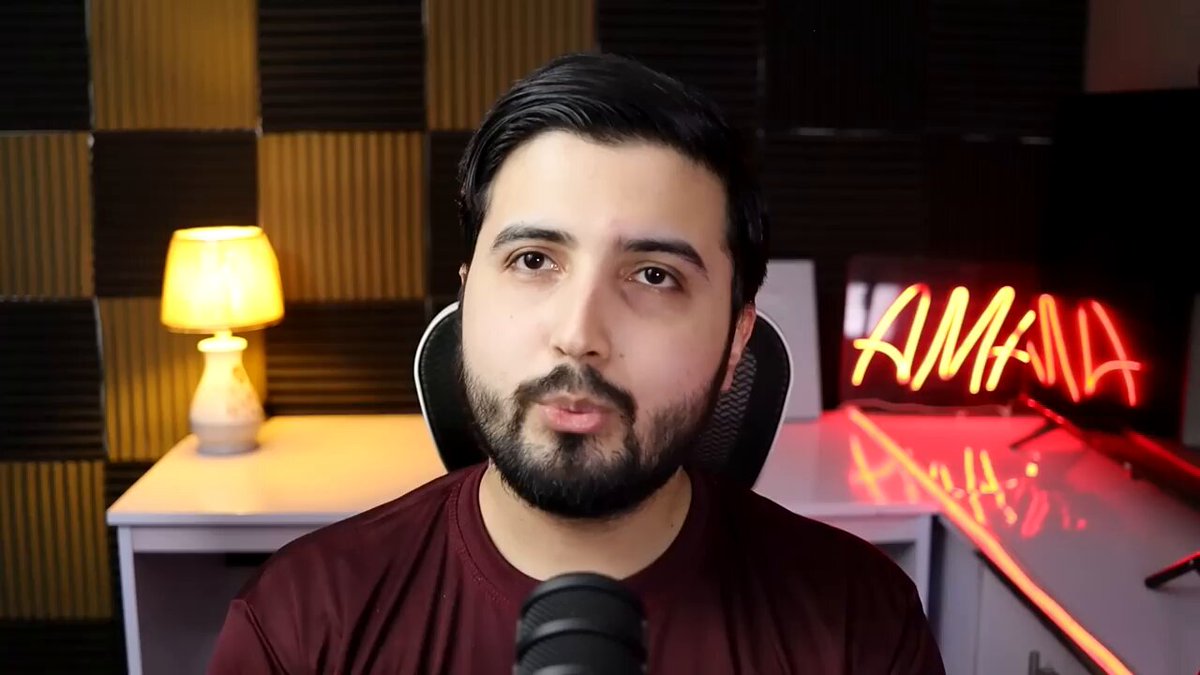 The image features a man with a beard wearing a red shirt sitting behind a microphone, likely recording or broadcasting something. The background includes a black wall with a neon sign that reads "Amana" written in white letters. There are also two lamps on either side of the man and a TV screen visible in the scene.