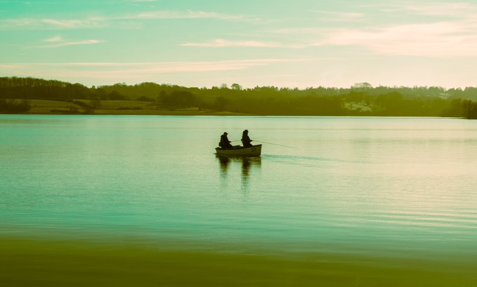 A scenic view of two people fishing from a small boat on a calm lake at sunset, with trees lining the distant shore.
