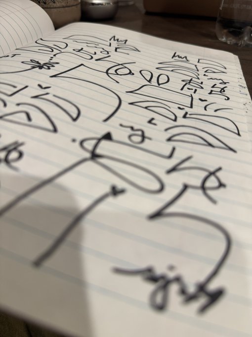 A close-up angled view of a lined notebook page filled with black handwritten text that appears to be in Japanese or Chinese characters, with part of another lined notebook visible at the top.