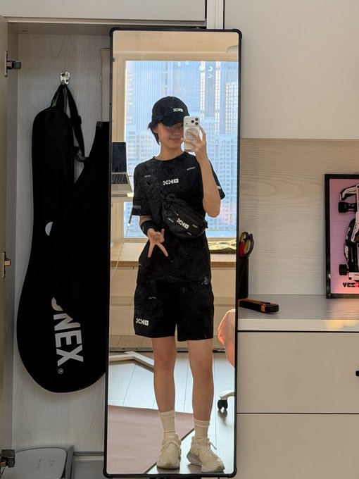 A person takes a mirror selfie wearing black JNEX athletic wear, cap, and fanny pack, with a JNEX branded bag hanging beside them.