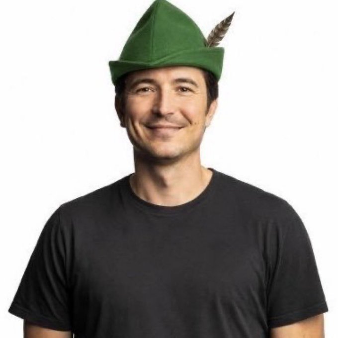 A man with short dark hair wearing a black t-shirt and a green felt Robin Hood-style hat with a feather, smiling at the camera against a white background.