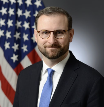 A man in a dark suit, blue tie, and glasses stands in front of an American flag, representing White House crypto adviser Patrick Witt as he prepares to take a months-long military leave amid critical negotiations over the Clarity Act.