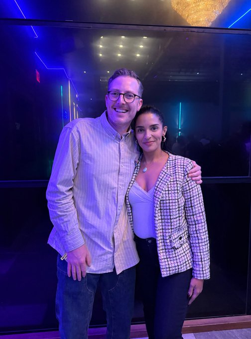 A man wearing glasses and a striped shirt stands with his arm around a woman wearing a pink checkered blazer, posing together in a venue with neon lighting and a chandelier.