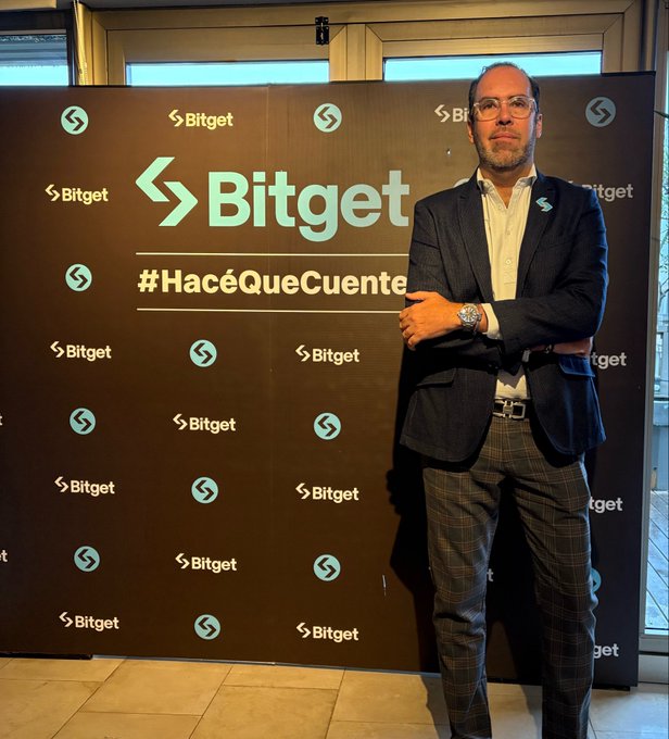 The image features a man standing next to a large black sign with white text that reads "BITGET". The man appears to be dressed professionally in a suit and tie, and he has his arms crossed over his chest. The background of the image shows a window, suggesting an indoor setting or possibly an event space where the sign is displayed for people to see. The post on the wall seems to be related to Bitcoin or cryptocurrency, as indicated by the text "Ready to talk about UEX, IPO Prime and all the benefits of @bitget".