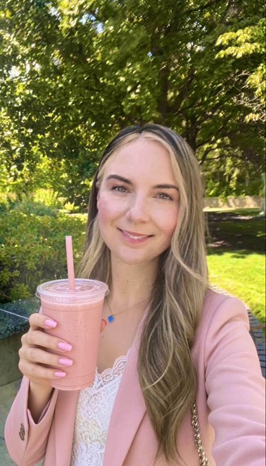 A young woman with long blonde hair smiles while holding a pink smoothie with a straw, wearing a pink blazer over a white lace top outdoors.