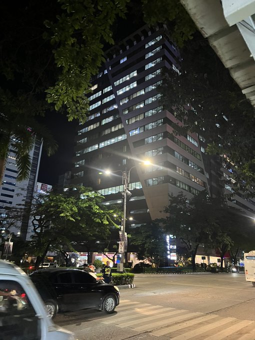 Nighttime street scene featuring a tall modern building with illuminated windows behind trees, with cars on the road and a streetlight glowing in the foreground.