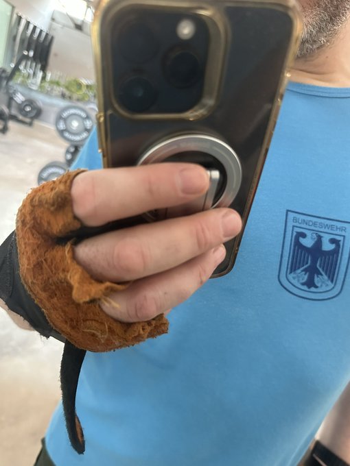 The image shows a man taking a selfie of himself with a black phone using his left hand. The man is wearing a blue t-shirt that has the emblem of the German Bundeswehr, indicating his affiliation or interest in the organization. The photo also features two additional words: "Double the amount! Two thousand !" and "Jumbo! Good luck Kameraden", which may possibly be an amusing phrase about the man's recent achievements at the Bundeswehr.