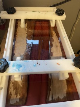A top-down view of a wooden project on a workbench clamped between wooden bars, showing wet resin or glue in blue and reddish-brown colors spread across the surface.