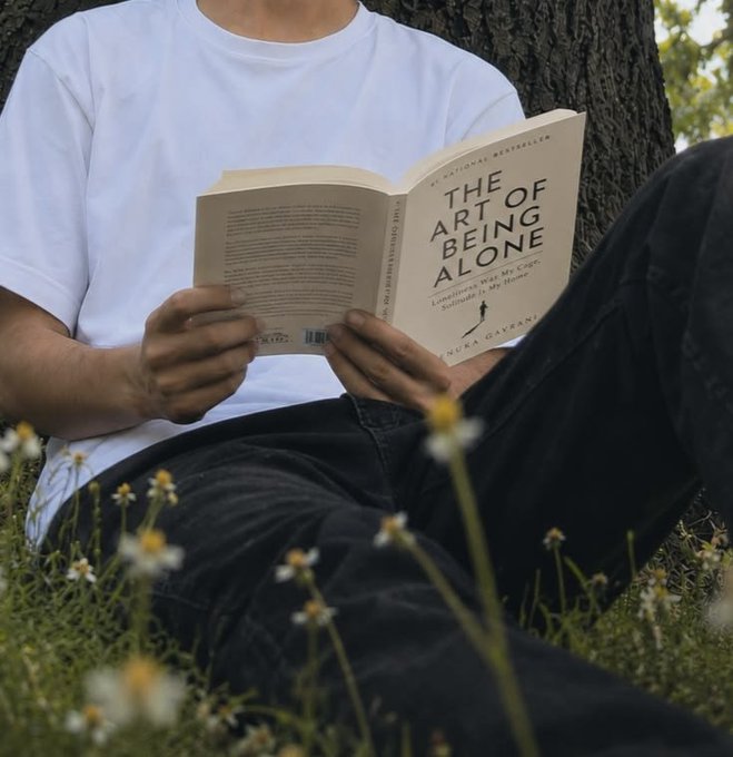 A person sitting outdoors against a tree reading the book "The Art of Being Alone" by Neha Garg.