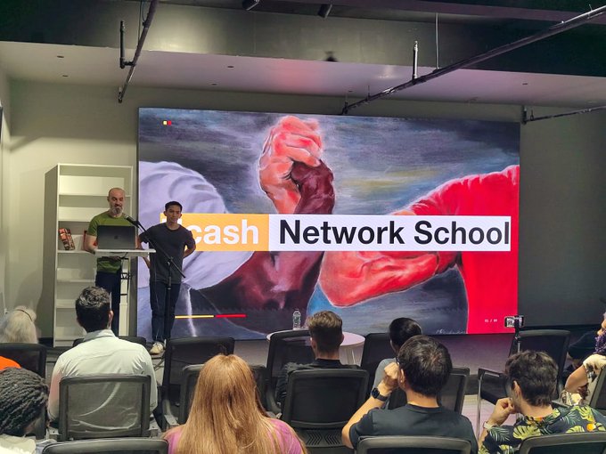Two presenters stand at a podium in front of a large screen displaying a painted mural of hands with the "Zcash Network School" logo, while an audience watches from rows of chairs below.

The image captures the closing event of the Zcash Network School, showing the final presentation to attendees after the successful completion of ZCG Grant #266.