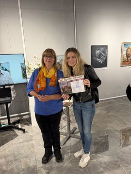 Two women stand in what appears to be an art gallery, one holding up a certificate or award while smiling for the photo.