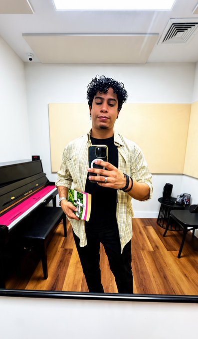 A young man with curly hair takes a mirror selfie with his phone while holding a mug in what appears to be a piano room with a black piano visible.