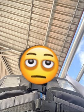 The image shows a low-angle selfie taken from what appears to be a backpack, with a tired/blank-face emoji covering the person's face. The caption "this user almost drowned today while touching grass" suggests a humorous contrast between the mundane outdoor setting (visible sky and overhead structure) and the dramatic "near-drowning" claim, implying a comedic exaggeration of a minor outdoor mishap.