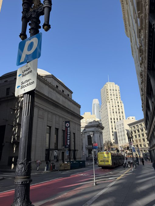 A street scene in San Francisco featuring a parking sign pointing toward Union Square and Sutter-Stockton, alongside a historic building, an ambulance, and skyscrapers under a clear blue sky.