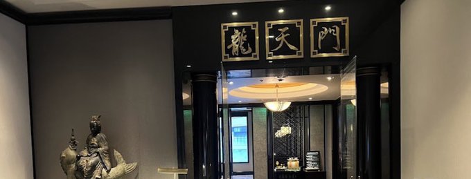 The entrance to a Chinese restaurant featuring gold characters '龍天門' (Dragon Heaven Gate) displayed on a dark wall above black pillars, with a statue visible to the left and a lit interior beyond.