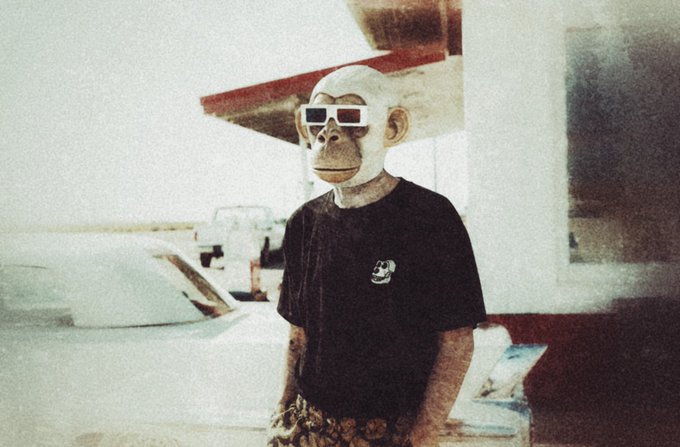 The image features a man wearing a monkey mask, sunglasses, and glasses. He appears to be standing outside of a building or an airplane hangar. There are two cars visible in the background, one parked near the left side of the image and another further back on the right side. The caption reads "GM bored ape yacht club", which suggests that the man's attire and location might be related to a themed event or gathering at the GM bored apes yacht club.