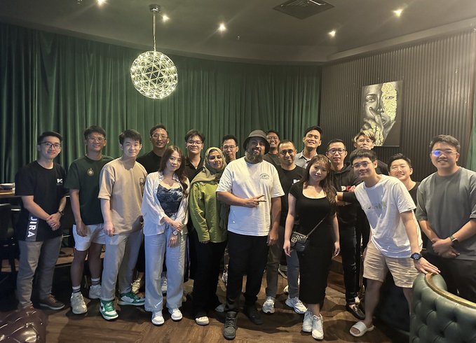 A diverse group of around 15 people poses together indoors with green curtains and a decorative spherical chandelier overhead, including a central figure in a white t-shirt and cap holding what appears to be an award or trophy. The group photo captures the IRL community spirit celebrated in the post, showing the KL meetup attendees who gathered in person to network—the kind of in-real-life connection the caption champions as "the alpha."