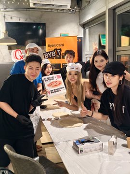 The image features a group of people posing for a photo around a dining table, with pizza boxes placed on top of the table. The individuals are wearing hats and appear to be enjoying themselves as they pose together. In the background, there is a TV screen mounted on the wall. The text in the image includes mentions of "MUShanghai", "bybit", " crypto markets", and "PizzaDay".