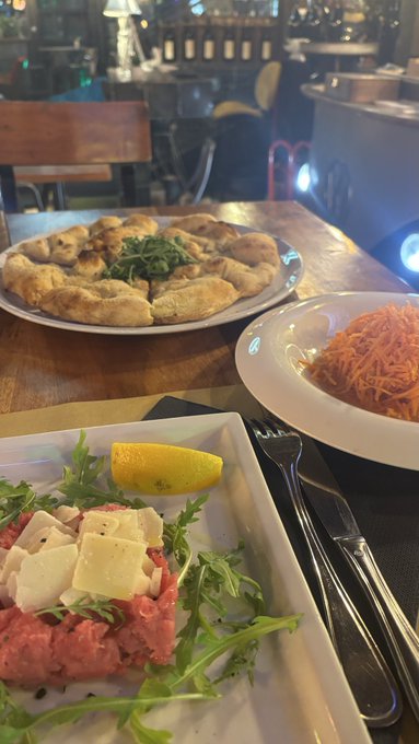 A restaurant table set with a plate of beef carpaccio with cheese shavings and arugula, alongside plates of bread, grated carrots, and a lemon wedge.