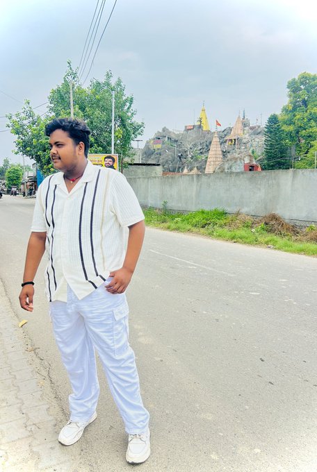 A man wearing a striped white shirt and white pants stands on a paved road, with a temple structure visible on a rocky hill behind him.