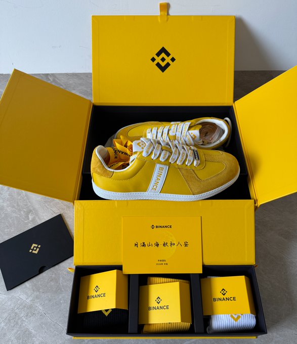 An open yellow Binance branded box containing a pair of yellow sneakers, a card, and rolled items, featuring the Binance logo and Chinese text.