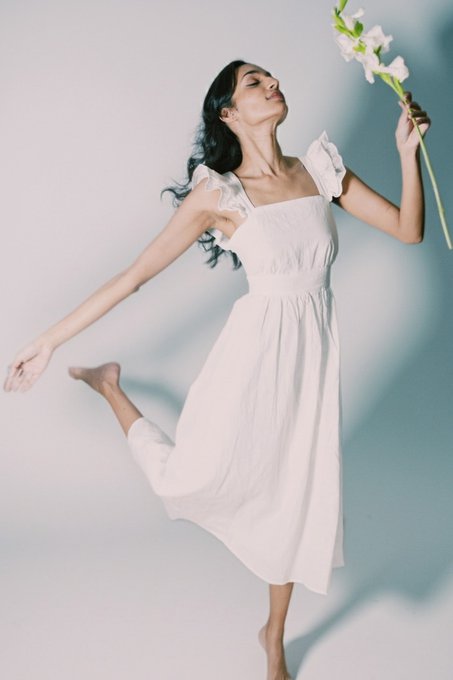 A woman with long dark hair dances barefoot in a flowing white dress, holding white flowers in one hand while balancing on her other leg against a soft blue-grey backdrop.