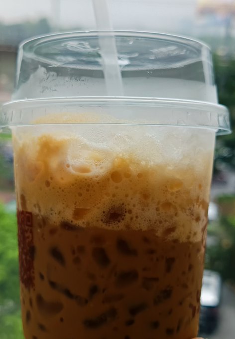 A close-up of an iced coffee drink in a clear plastic cup with a domed lid and straw, showing layers of coffee and foam.