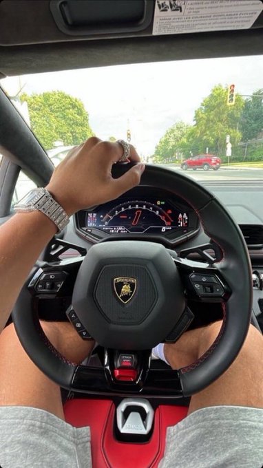 First-person view from inside a Lamborghini showing a hand with jewelry and a silver watch gripping the steering wheel at a traffic light.