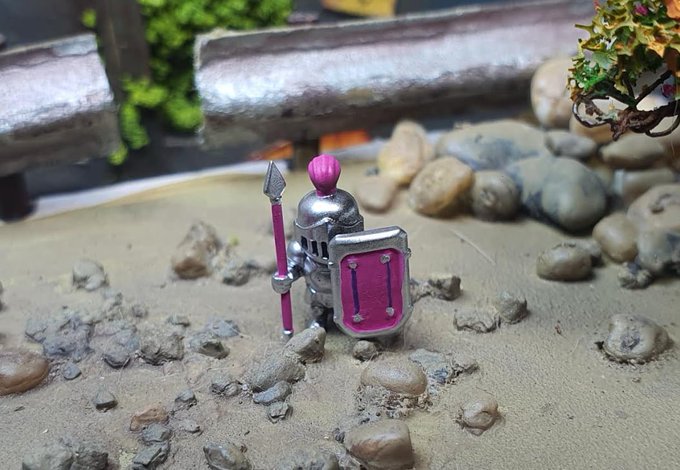 A miniature toy knight in silver armor with pink accents holds a spear and shield while standing on sand among small rocks near a miniature bridge.