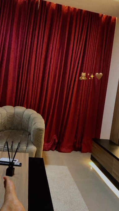An interior view of a room featuring rich red velvet curtains, a beige tufted sofa, a dark coffee table with a reed diffuser, and decorative bear and heart emojis.