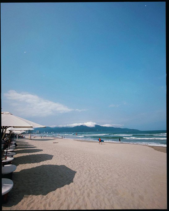The image shows a beach scene with people walking along the shoreline, some of them carrying umbrellas. There are several white umbrellas scattered across the sand, providing shade for those enjoying their time at the beach. The sky above is blue, indicating that it's daytime and perfect weather for outdoor activities like sunbathing or swimming in the ocean.
