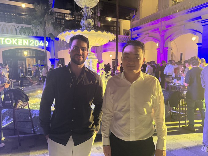 Two men pose together at the Token2049 speakers event at Raffles in Singapore, with the illuminated "TOKEN2049" signage and a grand tiered fountain visible in the background of the elegant venue.